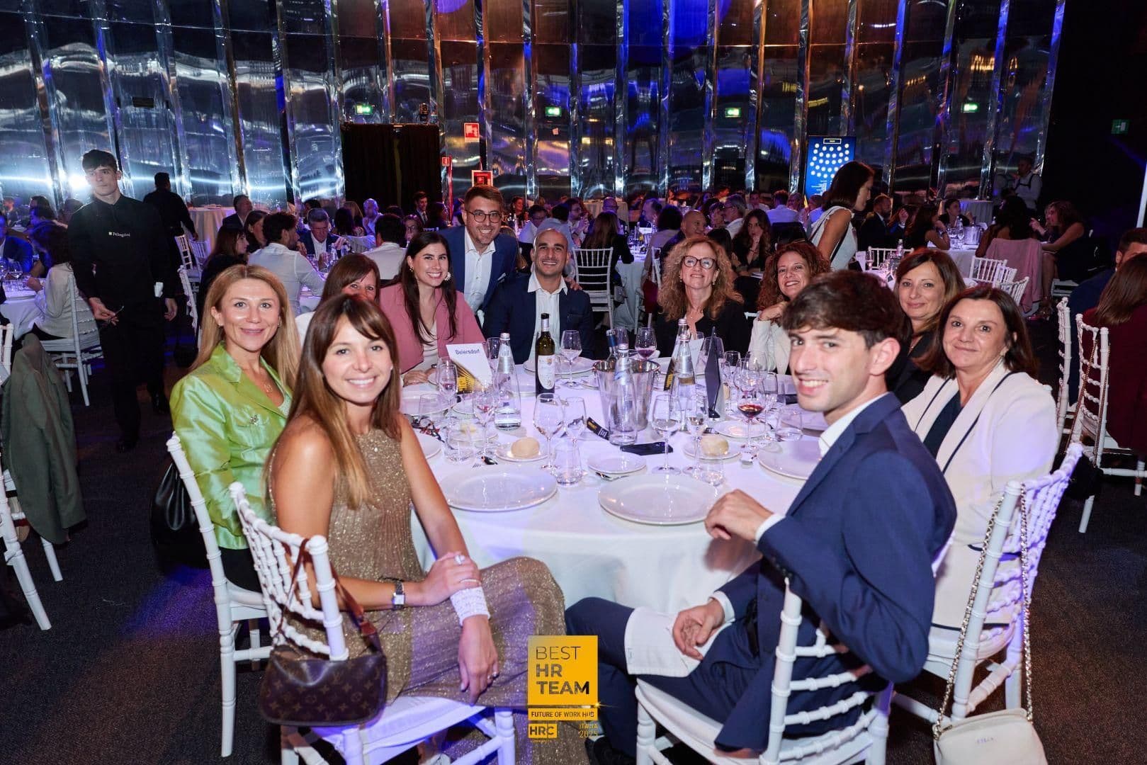 Donna e uomo sorridenti seduti a un tavolo elegante a una cena di gala, con piatti, bicchieri e altre persone sullo sfondo.