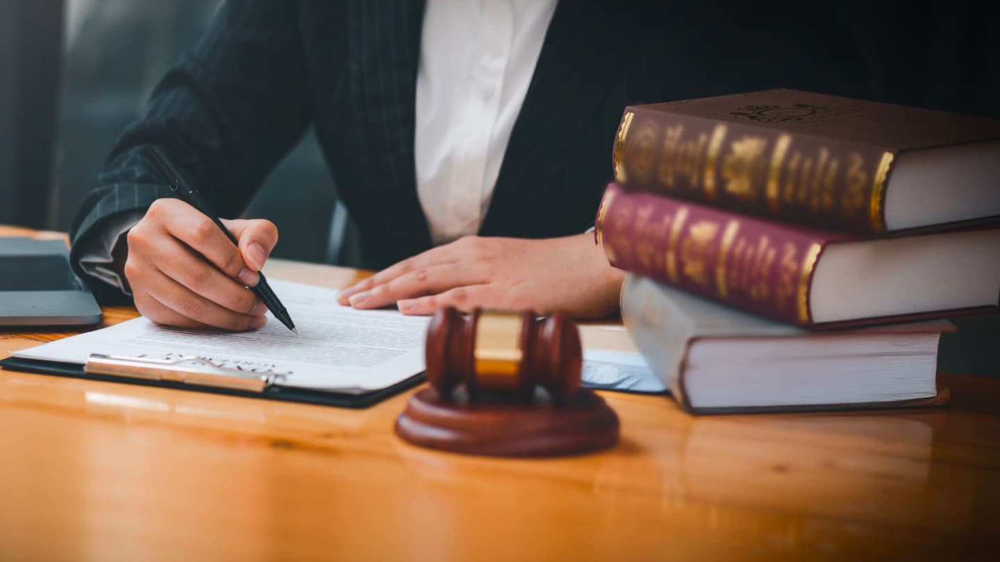 L’immagine mostra una persona in abito formale che scrive su un documento legale, con libri di diritto e martelletto da giudice sulla scrivania.